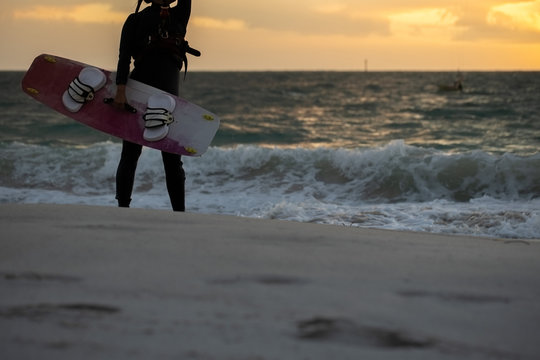 Picture of professional female kitesurfer wearing a black wetsuit standing holding up kitesurfing board equipment sideway prior surfing riding on the wave sunsetting on the beach at the background