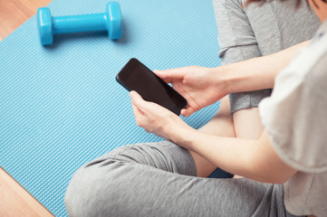 Top view of female person sitting in lotus position and looking at of smartphone screen. Isolation period. Copy space blank display of cellphone. Sports mat and dumbbell. Healthy life style concept