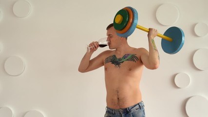 Young man with naked body and tattoo on chest lifting multicolored barbell and drinking red wine against white patterned wall. Adult strong guy doing sport and drinking alcohol.