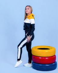 pretty young blonde model girl in sportswear posing next to car multi-colored tires in blue backround