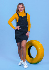 pretty young blonde model girl in sportswear posing next to car yellow tires in blue backround