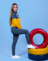 pretty young blonde model girl in sportswear posing next to car multi-colored tires in blue backround