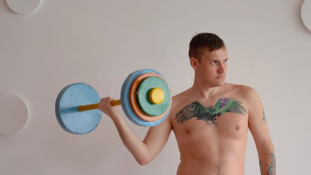 Young man with naked body and tattoo on chest lifting multicolored barbell against white patterned wall. Adult strong guy doing sport at home.