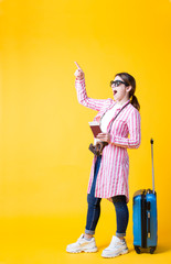 Emotional happy asian young woman tourist in stylish clothes standing with suitcase,pointing finger away on blank copy space for your advertising message text,promotional content on yellow