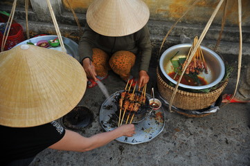 Street vendor with conical hat prepare roasted skewers on charcoal fire. Sidewalk barbecue is one...