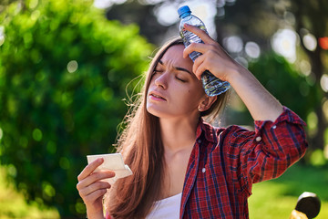 Tired sweating woman cooling with a bottle of cold refreshing water n a park in hot summer weather