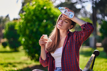 Sweating woman resting on a bench and cooling with a bottle of cold refreshing water in a park in...