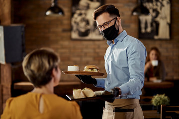 Happy waiter serving food to a guest while wearing protective face mask and gloves in a pub.