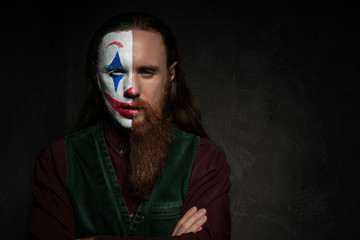 Man with long hair and beard and half face Joker makeup on dark background