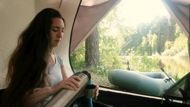Young woman tourist is sitting in a tent and drinking drinks from a thermos. Camping in the woods. Active lifestyle. Traveling in the morning. Camp by the river. A boat
