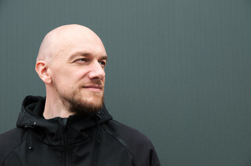 A young handsome bald man with a beard in the left part of the photo looks to the right, expresses confidence and determination. Dressed in a black jacket on a gray background. Place for text