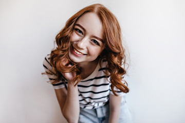 Indoor portrait of emotional ginger girl laughing to camera. Studio photo of excited caucasian lady wears striped shirt.
