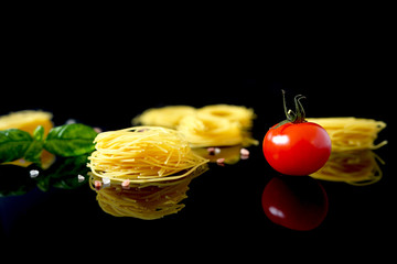 Dry, raw pasta spaghetti sockets with basil and red cherry tomato, salt. Reflection  on glossy black surface. Isolated.