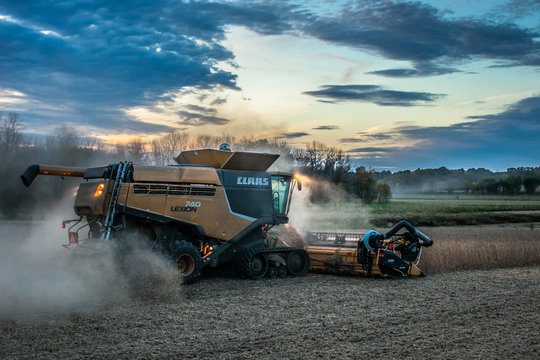 Side view of a Claas combine harvesting soybeans in the evening with the headlights on and dramatic skies.