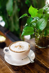 Cup of hot cappuccino and glass vase with green plant on wooden table in cafe.