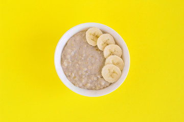 Bowl of oatmeal with banana on yellow background. Flat lay with copy space.