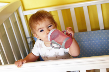 Cute one year old girl with blue eyes and blond is drinking water from bottle after waking up in...
