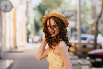 Laughing red-haired girl in hat posing on blur city background. Outdoor photo of winsome young woman with wavy hair having fun in summer.