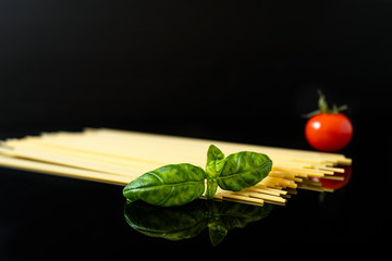 Raw spaghetti pasta and basil, red tomato, reflection of ingredients on a glossy black background, close-up.