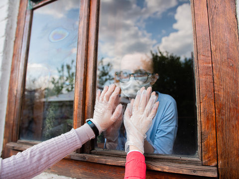A woman in a medical mask, leaning her hand against the window, communicates with relatives - Powered by Adobe