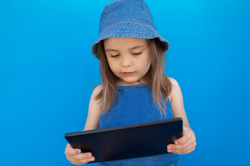 Portrait of a rather focused 5-year-old girl in a blue hat, against a blue wall and looking at a tablet.