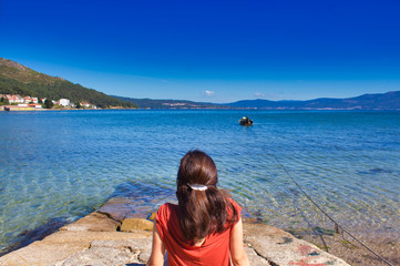 Fototapeta na wymiar girl on the port of Muros in Galicia, Spain having a relaxing time while she stares at the sea and a little boat
