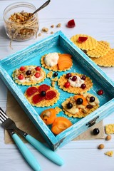 Waffles with berries and yogurt in a turquoise box, fork and spoon and a jar with granola on a light wooden background. Top view. Vertical
