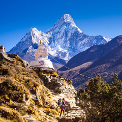 A woman with a large backpack, hiking on a rocky trail past a Buddhist Stupa with a snowy Ama Dablam peak behind.