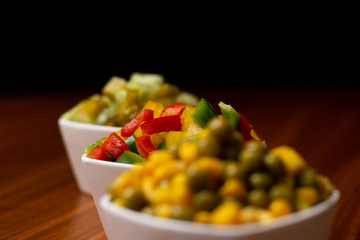 Papryki, ogórki, kukurydza i groszek pokrojone w kostkę do sałatki. Warzywa w białych miskach na brązowym stole gotowe do jedzenia. Świerze kolorowe warzywa na stole w białych miseczkach. 