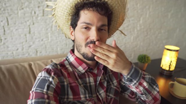 Cheerful Young adult brazilian man with straw hat having video call live stream and celebrating online Indoors at home living room. June Party holiday in Brazil, happiness, festival concept.