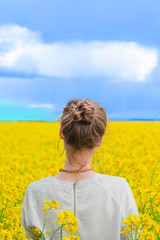 Fototapeta premium Brunette girl in rapeseed field summer time, back shot blue sky background. Happy beautiful young woman on blooming rapeseed field full of joy and happiness. Concept of freedom and childhood
