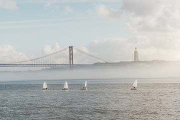 Fototapeta premium Foggy seascape of Tagus river in Lisbon, Portugal with four sailboats during a regatta sports competition or a performance, with a rope suspension bridge 25 Abril and statue of Jesus in the background