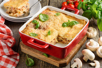 Lasagna with meat, mushrooms, tomatoes, cheese and bechamel sauce, decorated with basil in a red ceramic form. Traditional italian dish. Old wood background. Selective focus