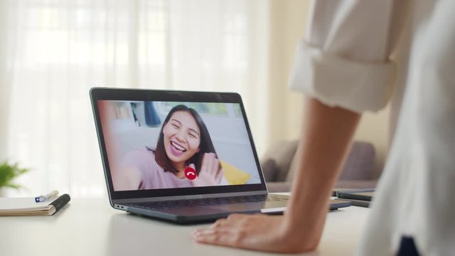 Young Asian business female using laptop video call talking with friends while working from home at living room. Self-isolation, social distancing, quarantine for coronavirus in next normal concept.