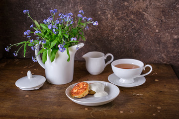 Fresh flowers of forget-me-not in a white kettle instead of a vase, a cup of tea, white milkman and a plate of cheesecakes made of cottage cheese with sour cream on an old wooden table.