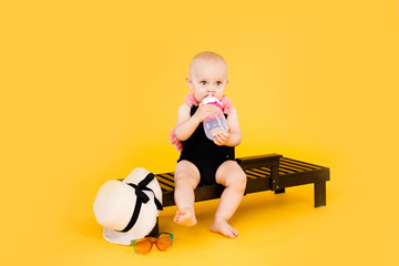 Funny little girl dressed in a black and pink swimsuit, big hat sitting on wooden deckchair with bottle of water on yellow background. Summer holidays concept