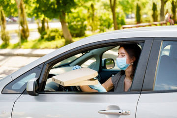 Student with pizza box in car with medical mask and gloves. Delivery food service drive by car. Safe food delivered according social distance. Tasty pizza with salami, cheese, tomatoes in cardoard box