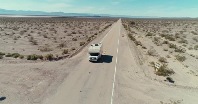 AERIAL - A drone turns around a small brown camper van that is driving in the Mojave desert on a sunny blue day