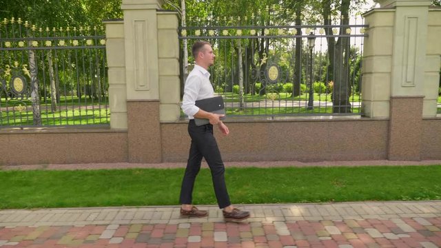 Businessman with laptop in his hand walks through the park. Man freelancer in a white shirt and trousers goes to a meeting with client. 4K footage