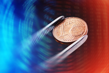 One euro cent in tweezers and financial data on background. Macro image.