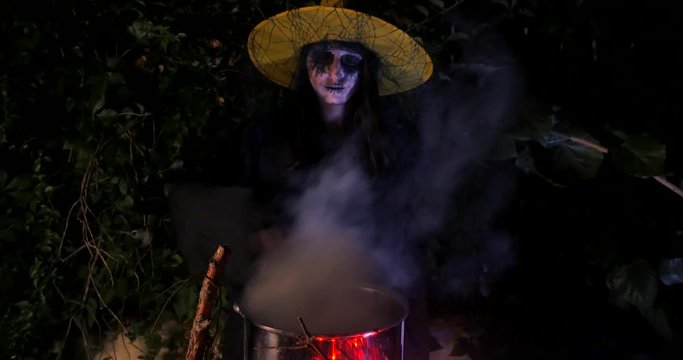Wicked frightening witch hexing with cane above the cauldron in dark forest . Halloween Horror Scene