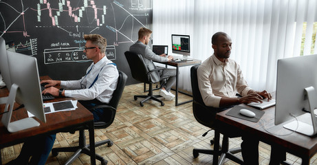 A world for learners. Three traders sitting by desks in front of computer monitors while working in the office. Blackboard full of charts and data analyses in background