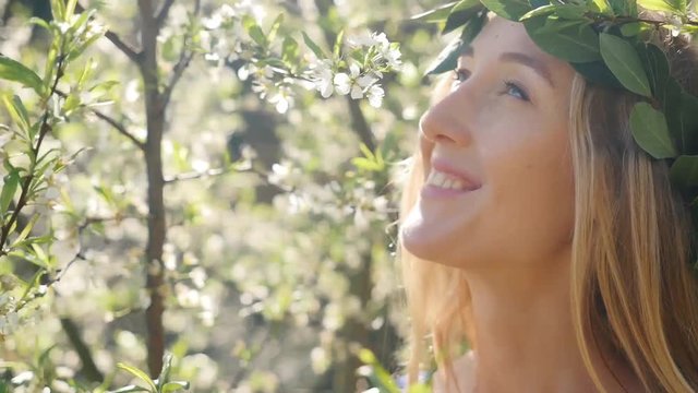 Portrait of smiling young beautiful woman with long blond hair in bay leaf wreath in spring blossom plum trees. Slow motion