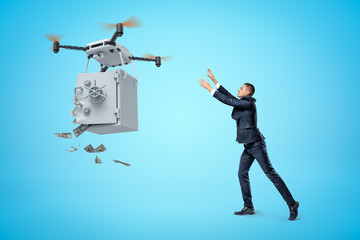 Businessman raising his hands to big quadcopter carrying money safe with open door and money falling out on blue background.