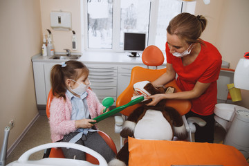 High angle of female dentist helping little patient to clean teeth of toy while working in modern clinic