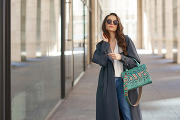 Stylish woman in white blouse and gray raincoat walking city street with reptile leather handbag....