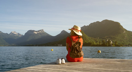 Girl with a stalk hat on and red dress looking at the mountains with her shoes on a dock by a lake