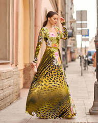 Pretty elegant female model wearing long colorful evening dress, standing and posing at city street...