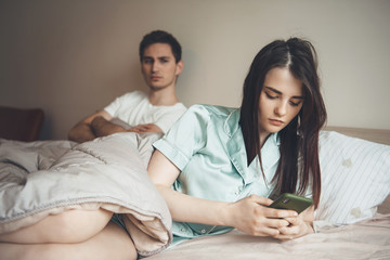 Worried caucasian man looking at his girlfriend chatting in bed on mobile