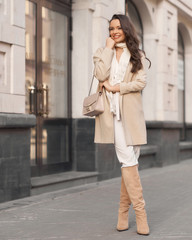Obraz premium Full length street portrait. Young elegant woman wearing beige stylish look. Pretty girl with long brunette wavy hair. Fashionable lady in white blouse, beige jacket, boots, scarf holding handbag